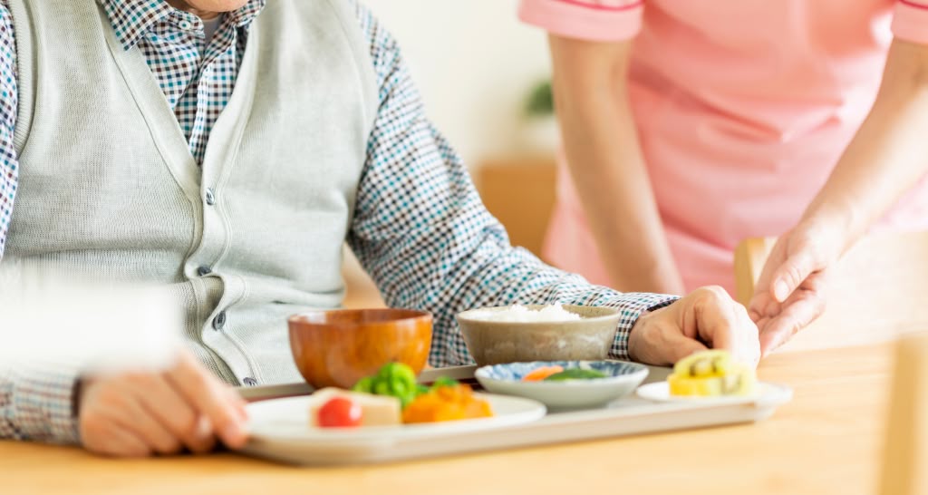 Pessoa idosa sendo assistida durante refeição, com pratos adaptados sobre a mesa.