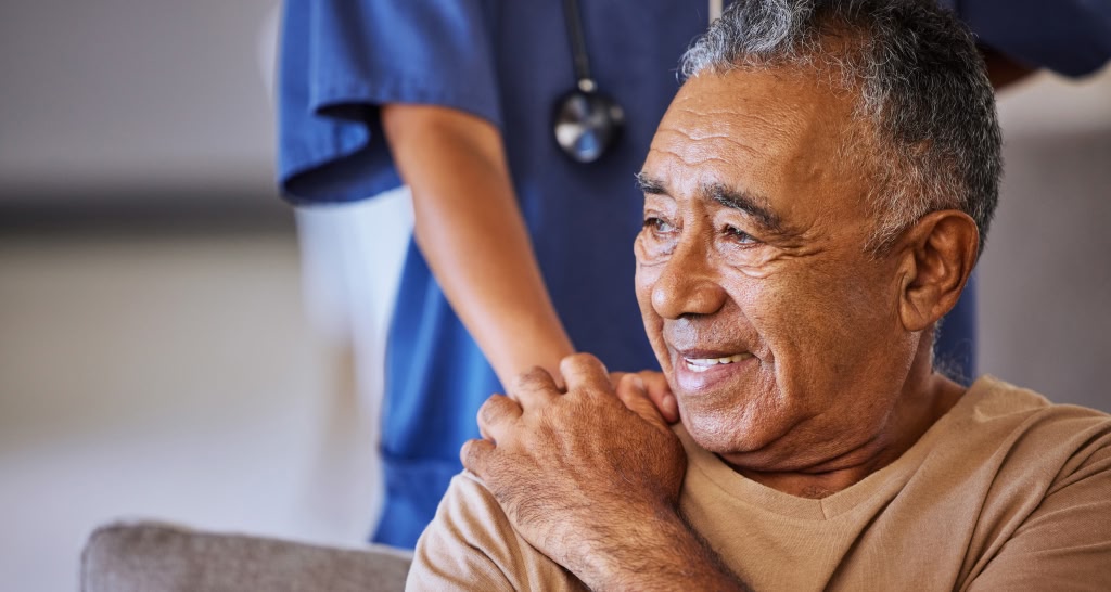 Profissional de saúde de jaleco azul e estetoscópio coloca a mão no ombro de um paciente idoso sorridente, transmitindo cuidado e acolhimento